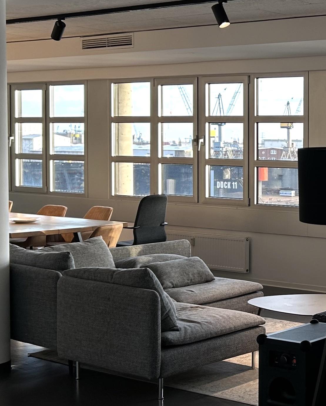 Offener Lounge- und Arbeitsbereich der Reneo Group in Hamburg mit Sitzlandschaft, Konferenztisch und Hafenkränen vor den Fenstern