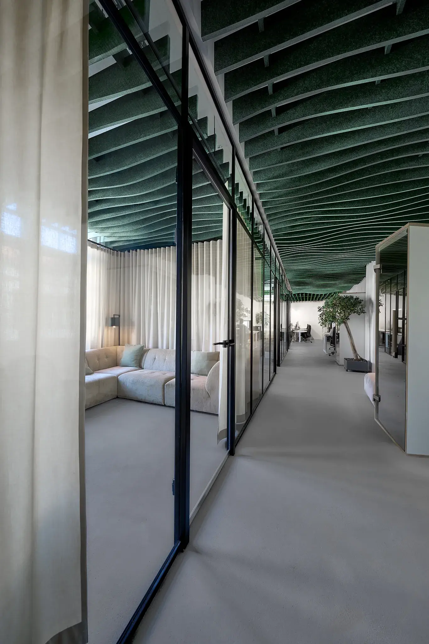 Moderner Büroflur mit schwarzen Stahlrahmen-Glaswänden, grüner Deckenverkleidung und Blick auf einen Loungebereich am Ende des Gangs.