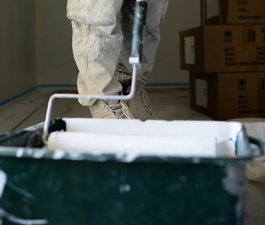 Nahaufnahme einer Hand mit Malerrolle, die weiße Farbe aus einem dunklen Farbeimer aufträgt.