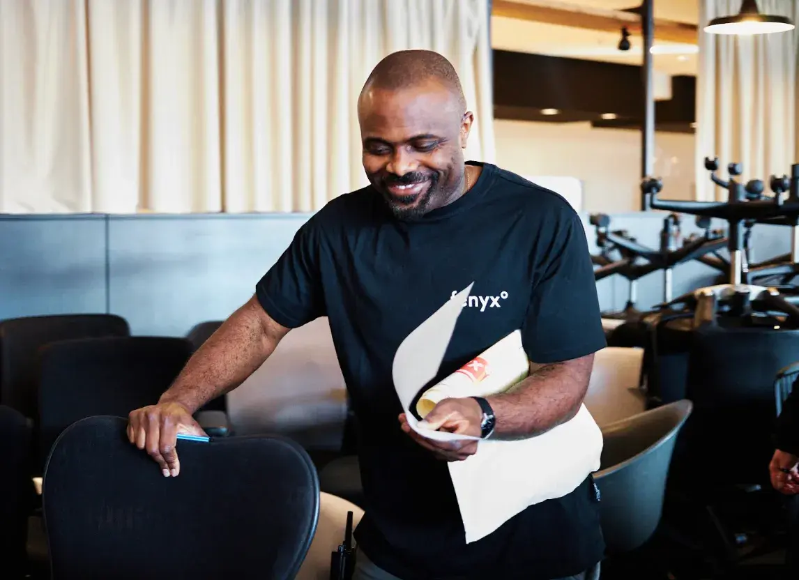Lächelnder Mann in schwarzem T-Shirt, der in einem modernen Lounge-Bereich sitzt und Papiere in den Händen hält.
