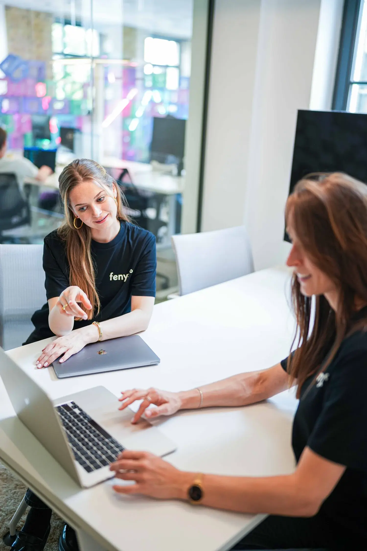 Zwei Frauen, die an einem weißen Tisch mit Laptops in einem modernen Büro zusammenarbeiten.
