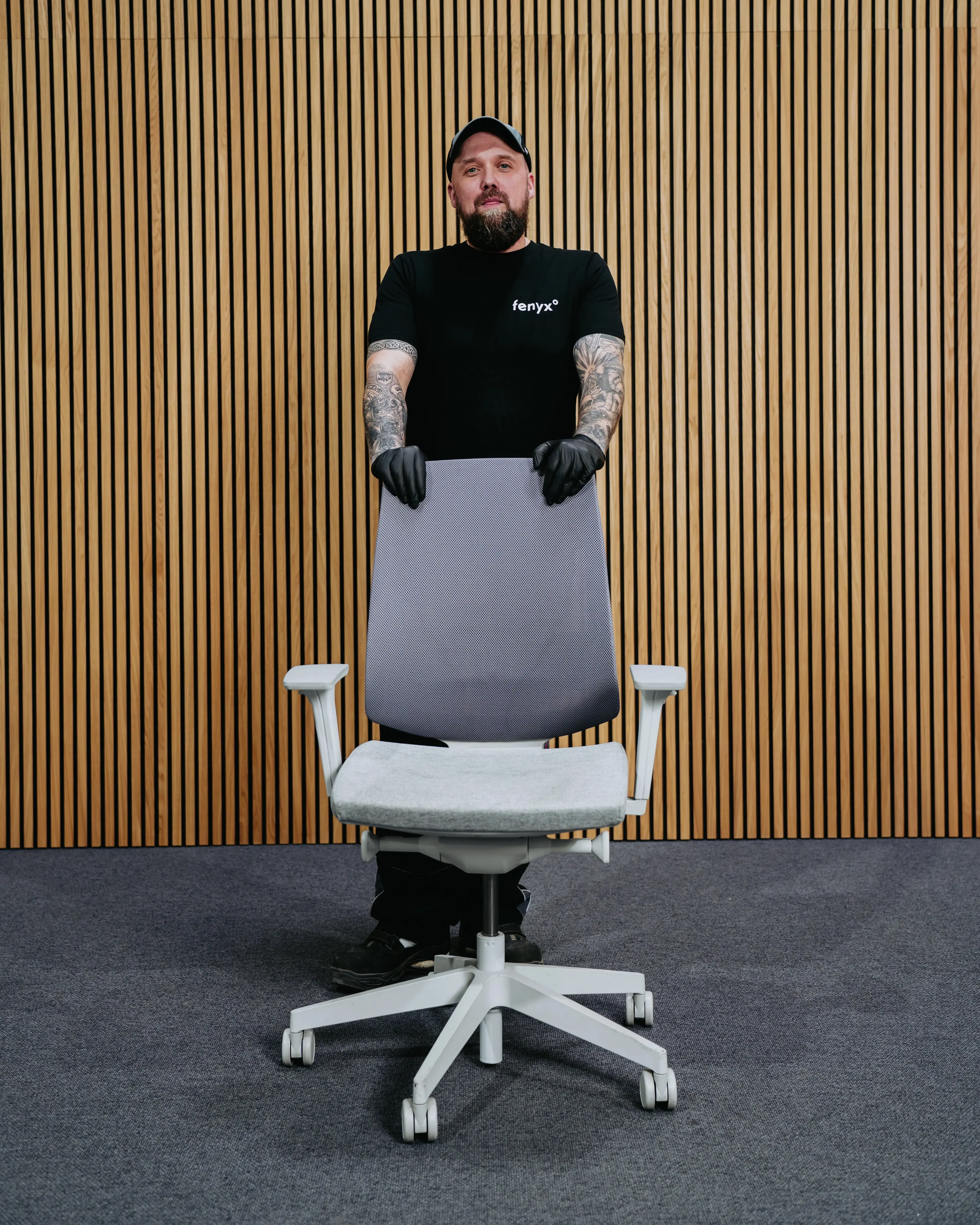 Mann mit Tattoos in schwarzen Handschuhen und schwarzem Shirt, der hinter einem grauen Bürostuhl vor einem Holzpaneelwand-Hintergrund steht.