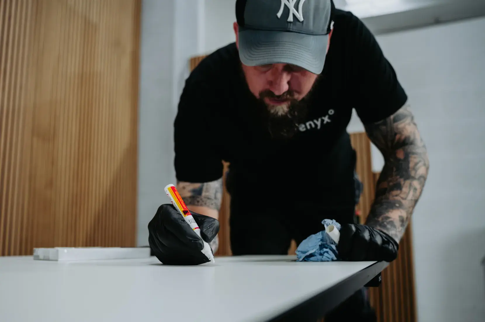Mann mit Tattoos und schwarzen Handschuhen, der Klebstoff auf eine weiße Oberfläche aufträgt und dabei ein blaues Tuch hält.