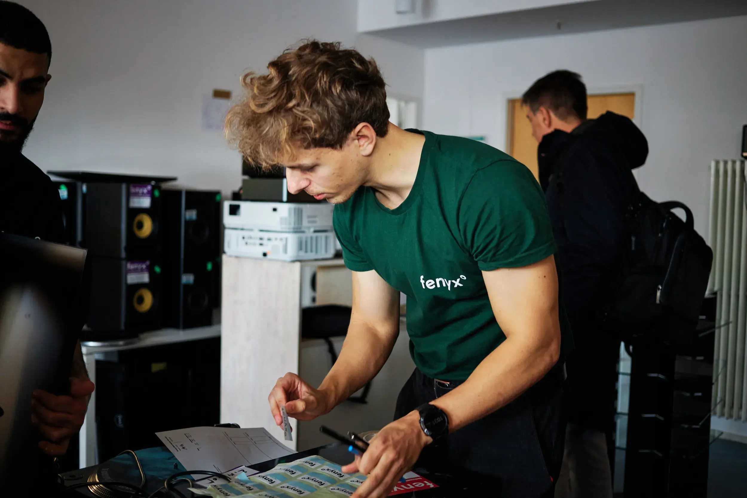 Junger Mann in einem grünen T-Shirt mit dem Aufdruck „fenyx“, der sich über einen Tisch mit Aufklebern und Papieren beugt und mit einer anderen Person interagiert.