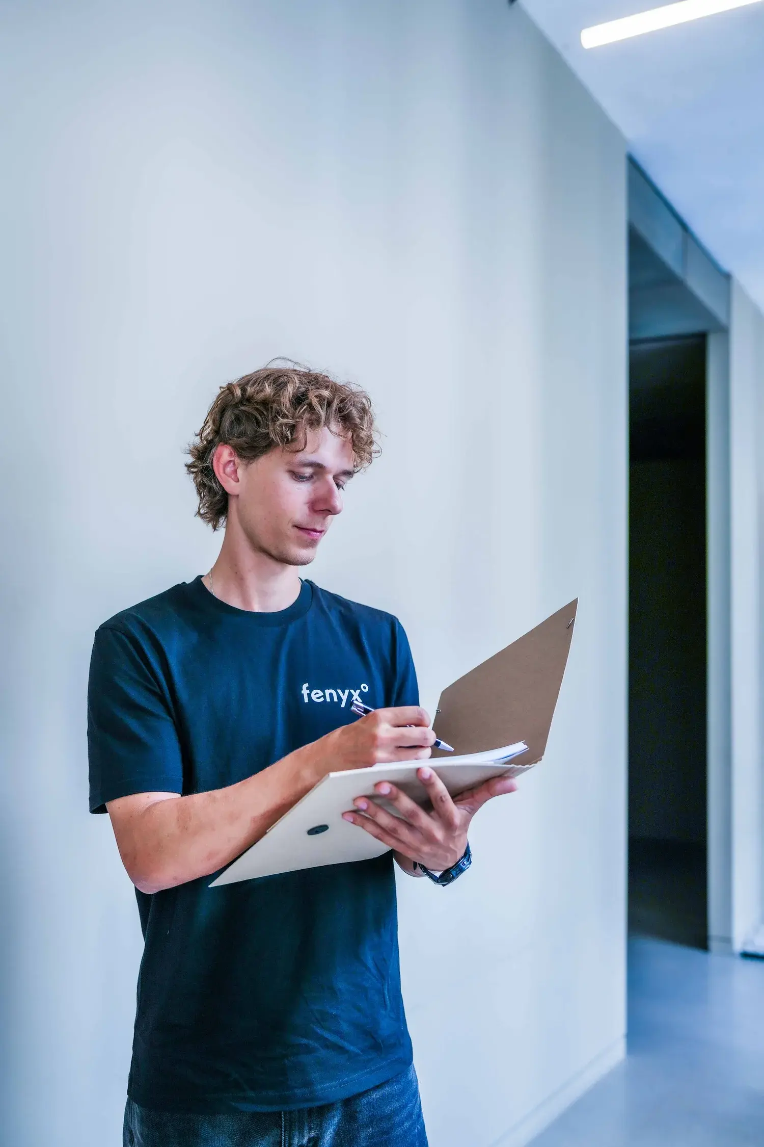Junger Mann mit lockigem Haar in einem dunkelblauen Fenyx-T-Shirt, der in einem hellen Flur auf einem Klemmbrett schreibt.