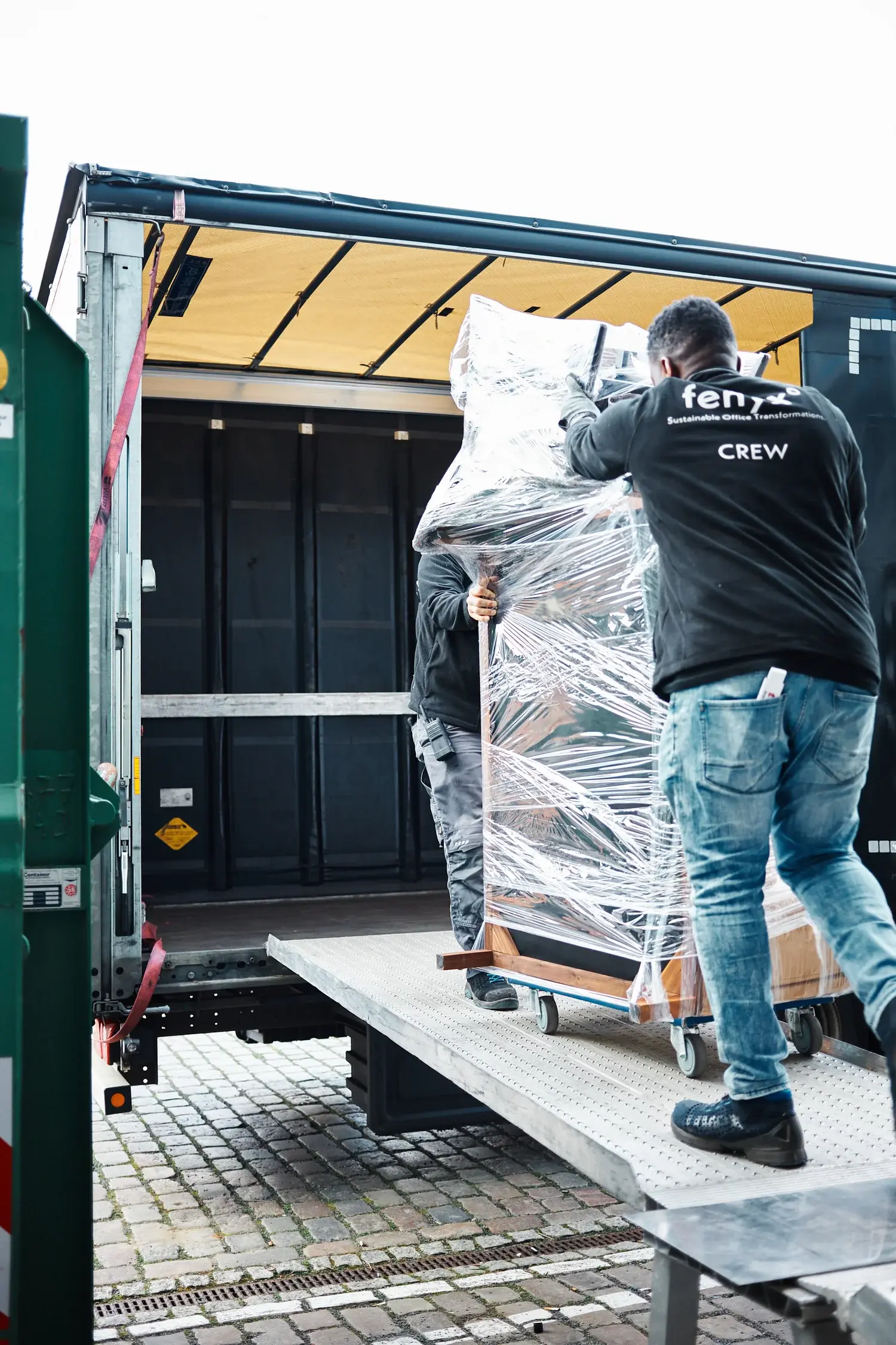 Zwei Crewmitglieder, die einen großen verpackten Gegenstand auf Rollen über eine Metallrampe auf Kopfsteinpflaster in einen Lastwagen laden.